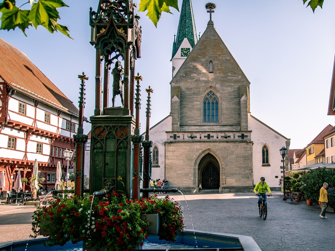 Historischer Marktplatz von Bad Saulgau mit gotischer Kirche, einem verzierten Brunnen mit Blumen sowie traditionellen und modernen Gebäuden; Menschen flanieren und ein Radfahrer fährt vorbei, bei sonnigem, Herbst-Wetter.